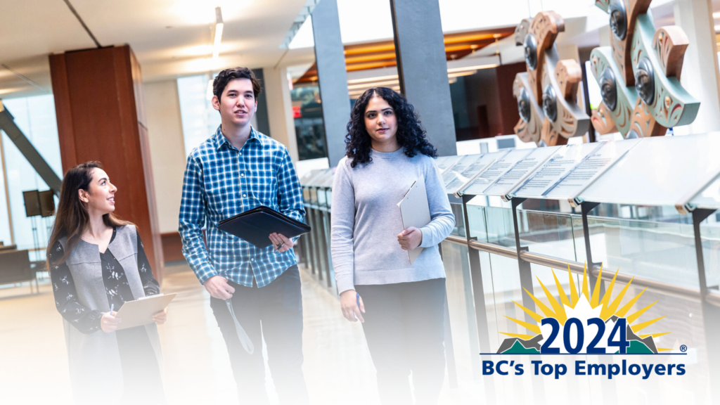 Three UBC staff members walking in the Henry Angus Building, each carrying a clipboard. On the bottom left of the photo is the logo for the 2024 BC Top Employers program.