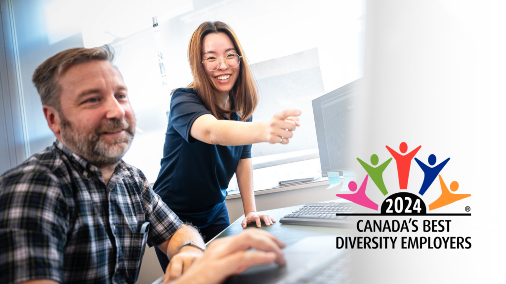 A picture of a seated bearded white man beside a standing Asian woman looking at a screen (off-camera). The woman is pointing at the screen. The logo for the 2024 Canada's Best Diversity Employers is superimposed over the bottom right of the photograph. 